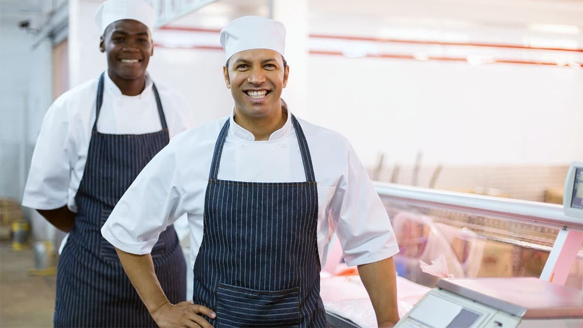 Two smiling employees wearing white uniforms and aprons, standing next to a scale